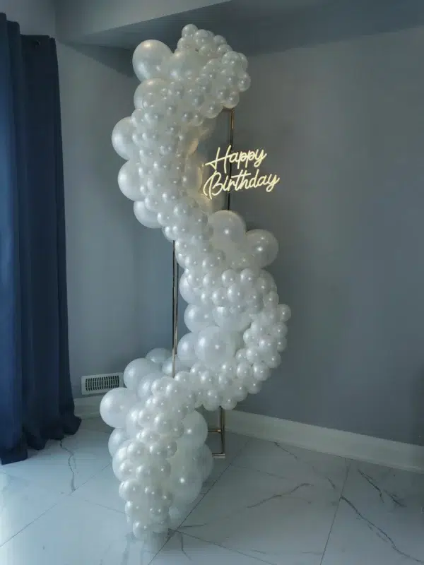 A decorative balloon arch in white, with a glowing "Happy Birthday" sign, set against a light blue wall. Perfect for birthday celebrations.