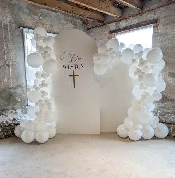 White balloon arch with a backdrop reading "God Bless Weston" and a gold cross, set in a rustic interior space.