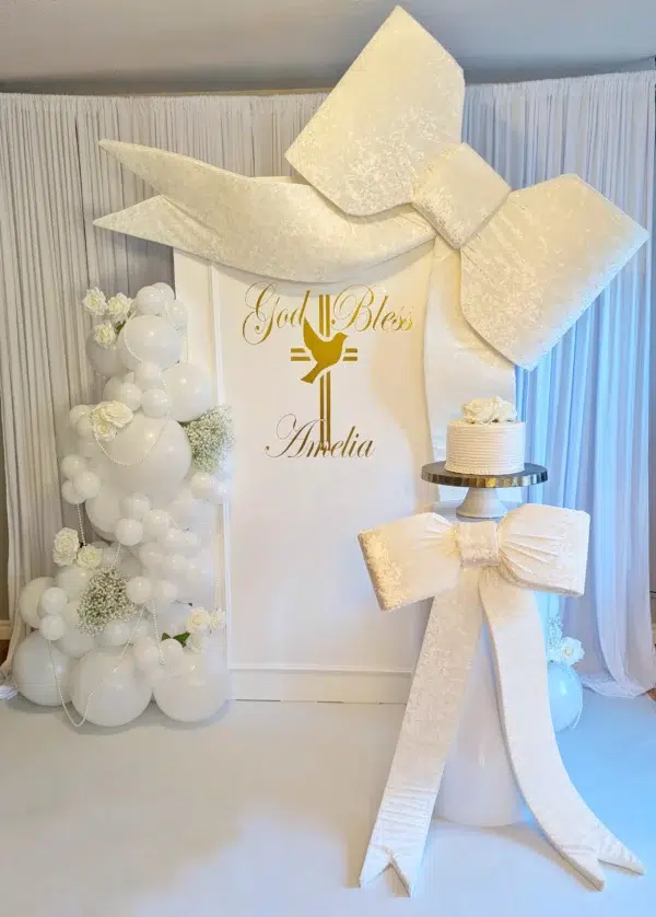 Elegant decor featuring a large white bow and balloon arrangements for a celebration. Gold lettering reads "God Bless Amelia."