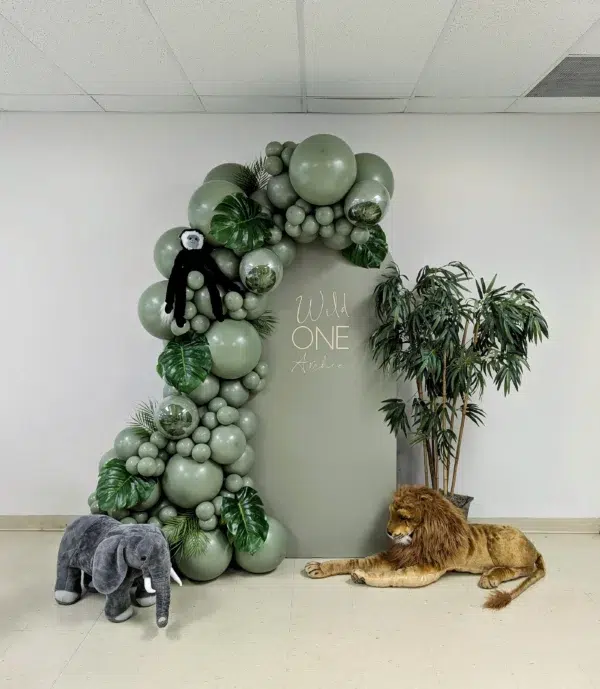 A whimsical party backdrop with green balloons, a plush monkey, elephant, and lion, featuring "Wild One" text for a jungle-themed celebration.