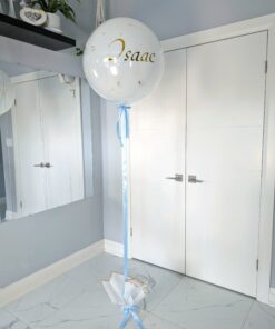 A large white helium balloon with gold foil specks and the name “Łukasz” in gold script, tied with a light blue ribbon to a decorative white fabric balloon weight with gold trim, displayed indoors against a light grey wall and white doors.
