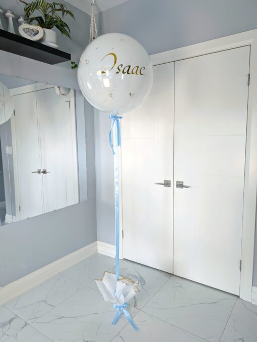 A large white helium balloon with gold foil specks and the name “Łukasz” in gold script, tied with a light blue ribbon to a decorative white fabric balloon weight with gold trim, displayed indoors against a light grey wall and white doors.