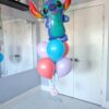 Stitch-themed balloon bouquet featuring a large colourful Stitch foil balloon and a cluster of pastel latex helium balloons with a small flower accent and a simple balloon weight.