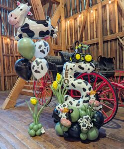 Farm themed balloon setup with cow print number stack, helium bouquet, greenery accents, and personalized name decal in barn setting