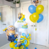 Minions-themed balloon setup with blue and yellow number stack, banana accents, helium bouquet, and personalized name decal