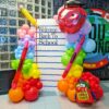 Rainbow back-to-school balloon display with a custom welcome backdrop, pencil, crayon, apple and backpack balloons.