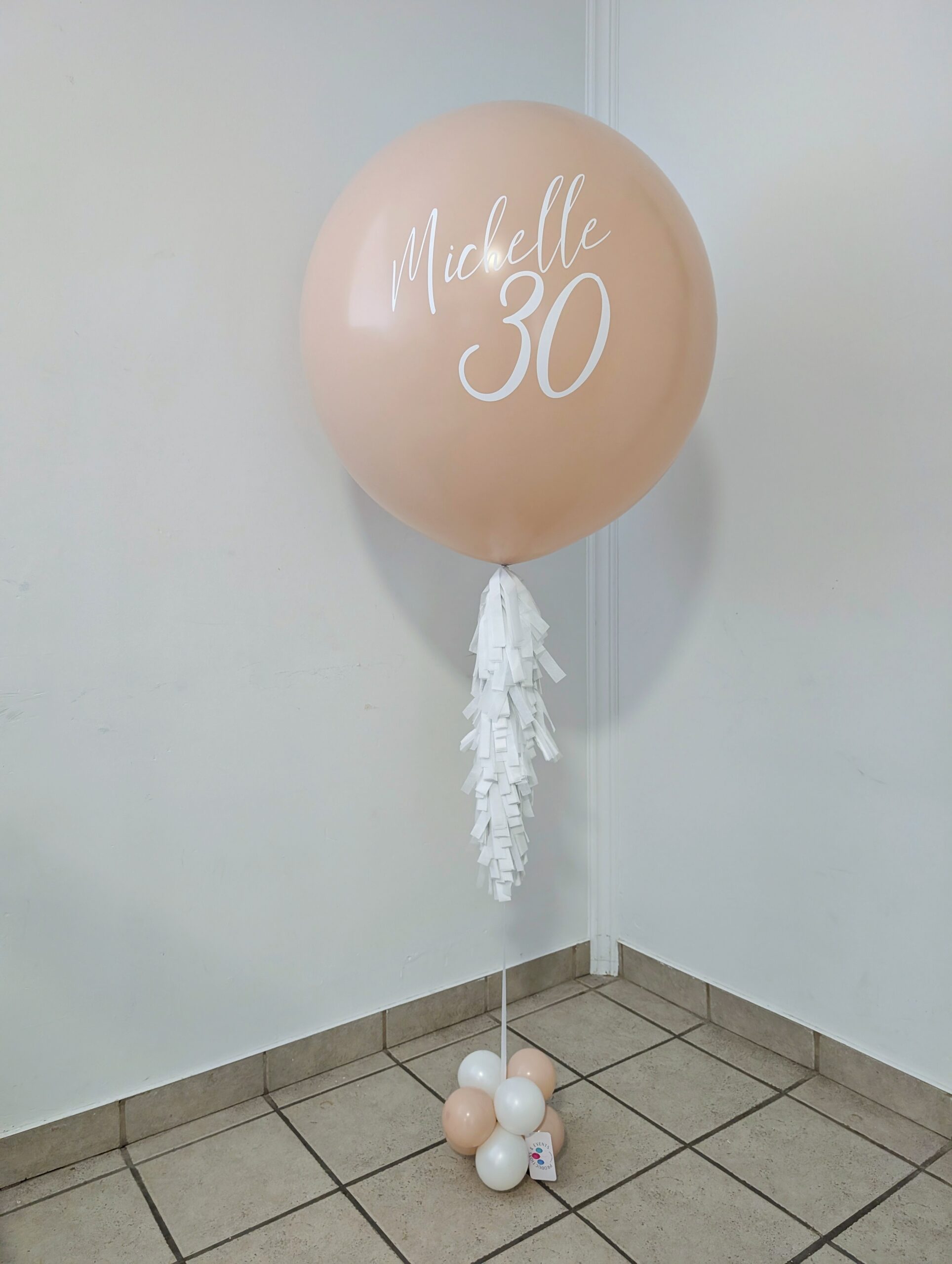 Personalized blush jumbo helium balloon with white lettering, tassels, and a coordinating balloon base.