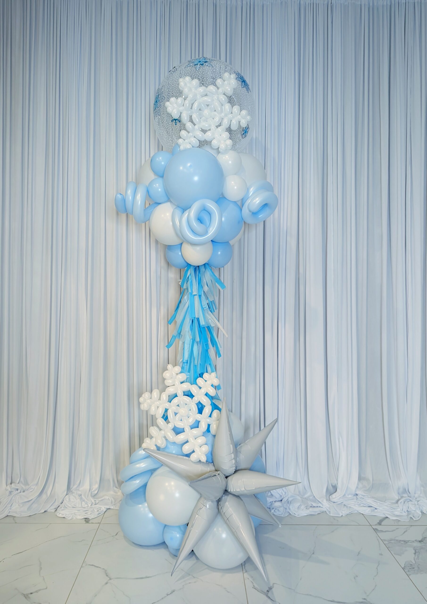 Blue and white winter balloon column with snowflakes, silver starburst balloons, fringe and a clear bubble topper.