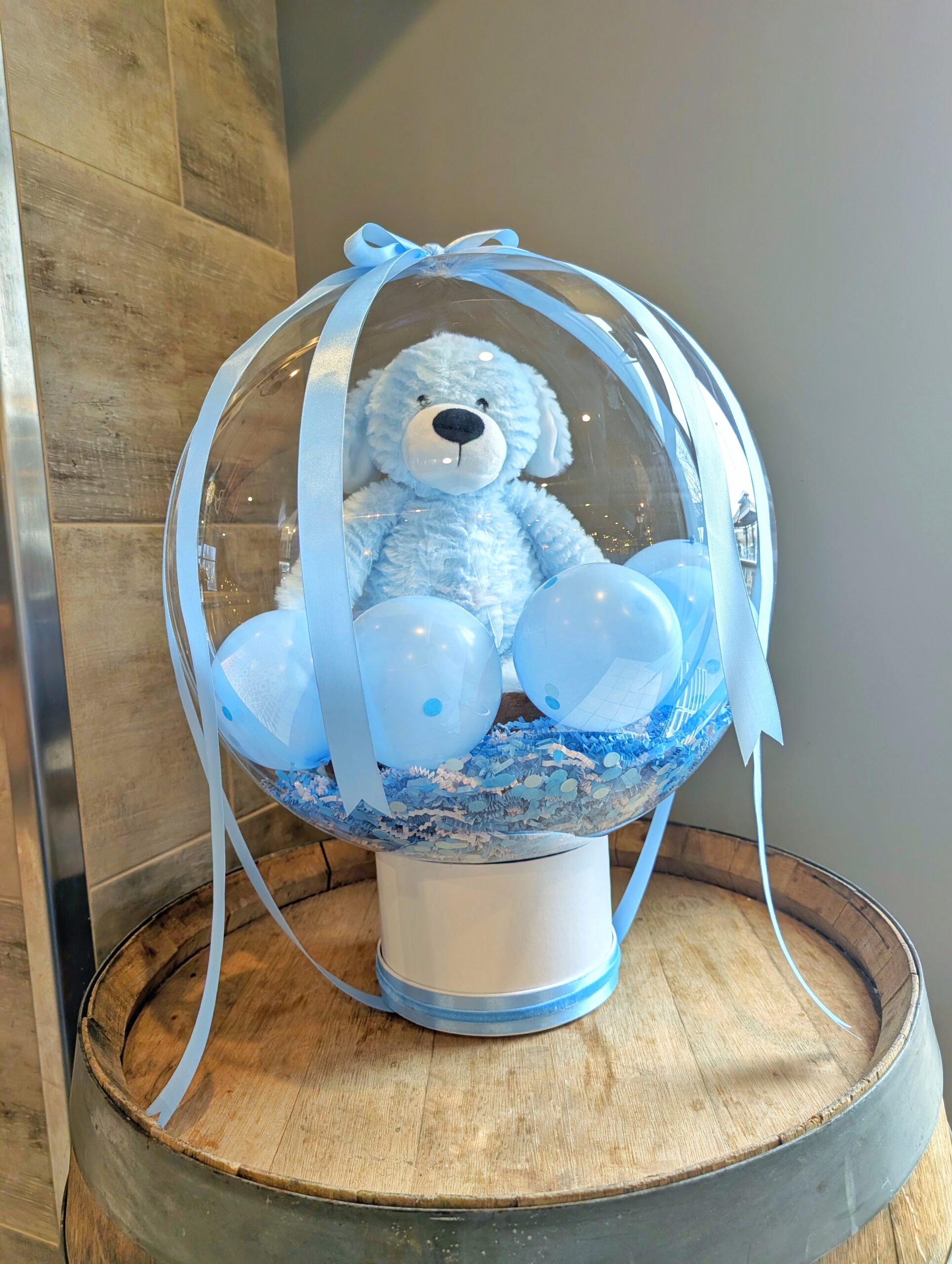 Clear stuffed balloon with a blue teddy bear, mini balloons, decorative shred, blue ribbons and a white base.