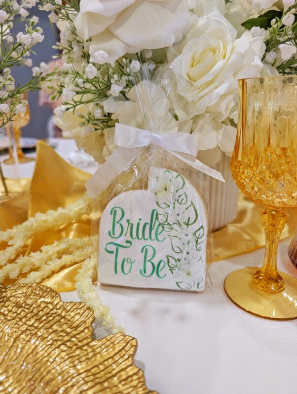 Bride-to-be table detailed sugar cookie beside white faux flowers, textured gold charger and amber glass