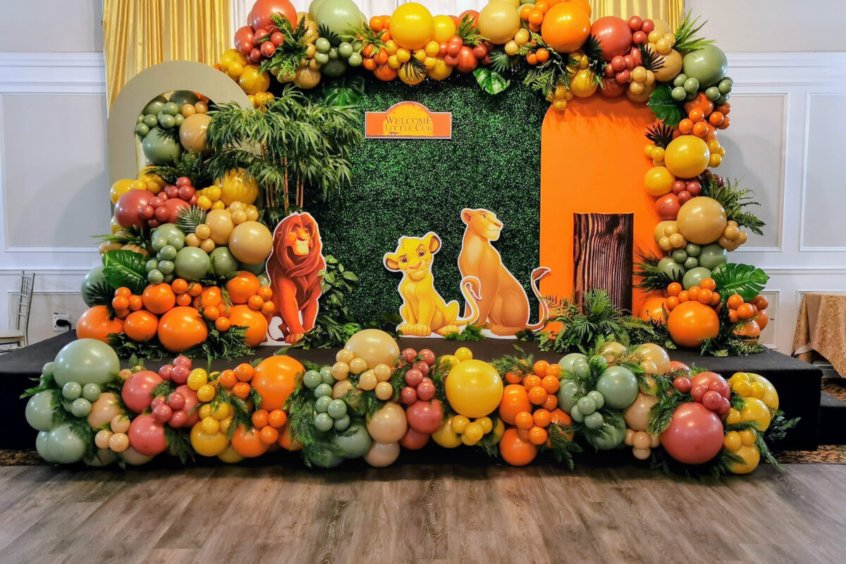 Lion King baby shower backdrop with earthy balloons, greenery, orange panels and character cutouts
