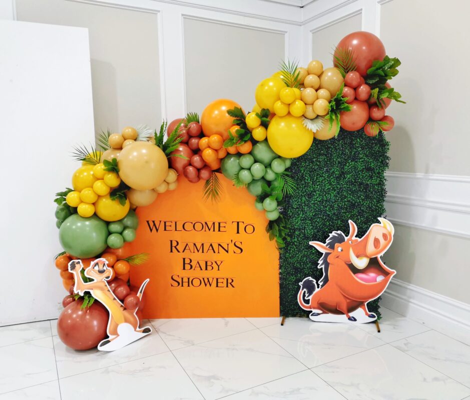 Lion King baby shower welcome display with orange panel, boxwood wall, balloons, Timon and Pumbaa