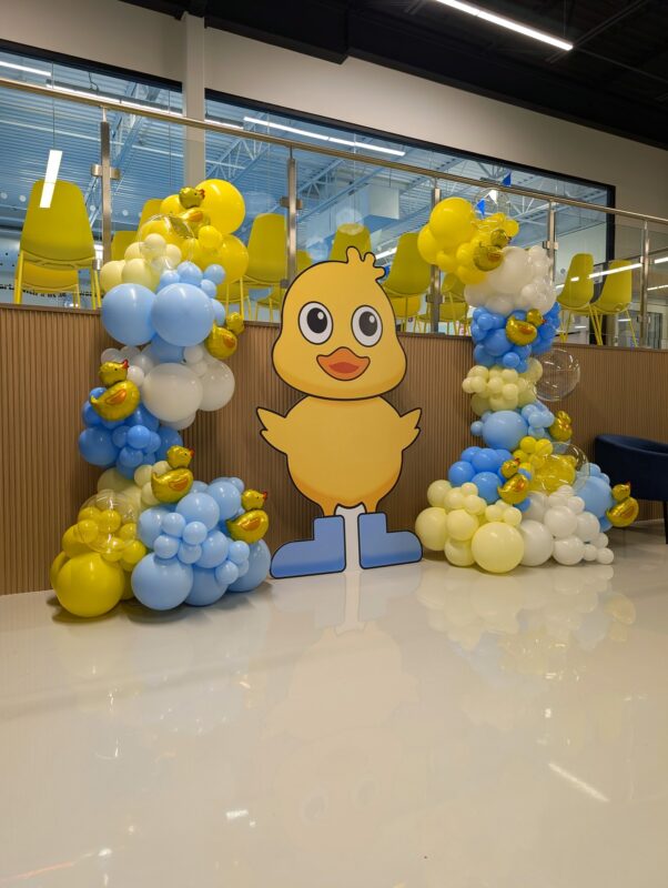 Custom rubber duck cutout framed by balloon columns and small duck-shaped foil balloons.