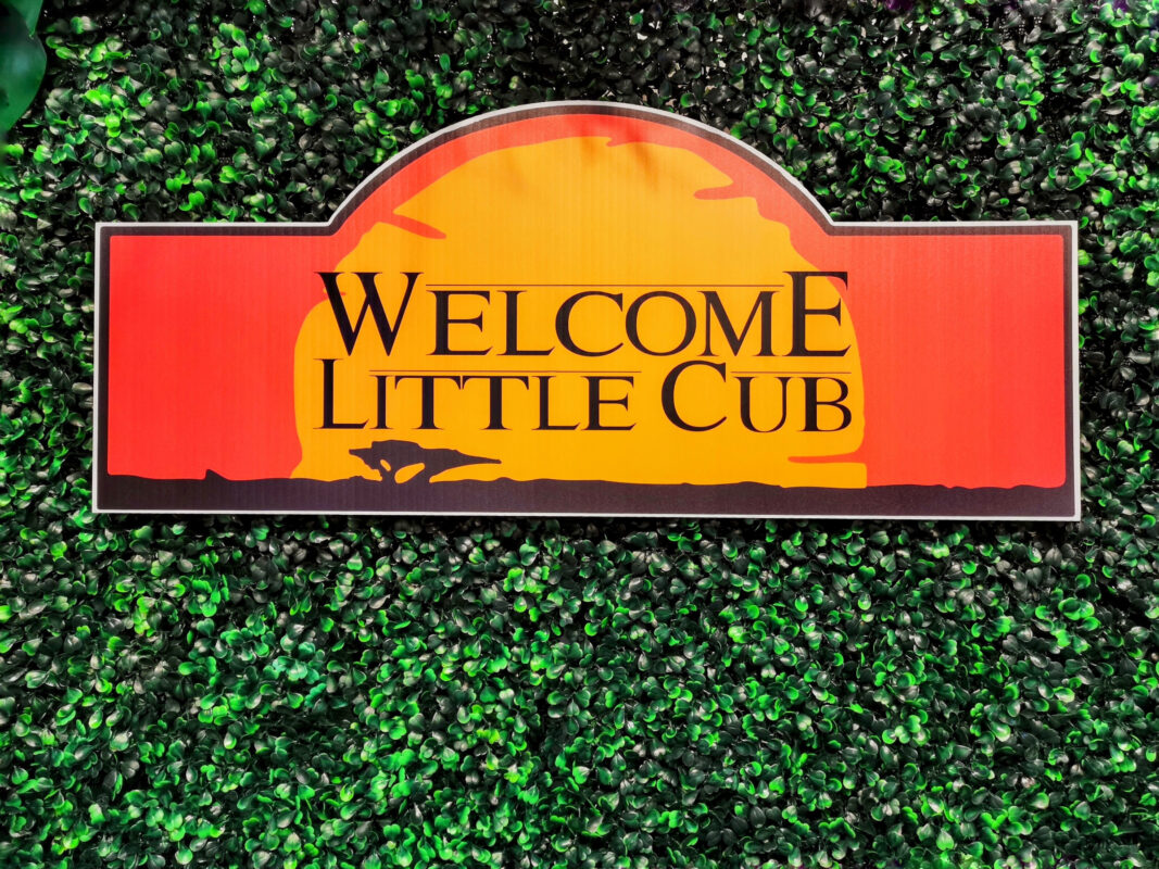 Custom Welcome Little Cub sign mounted on a green boxwood baby shower backdrop